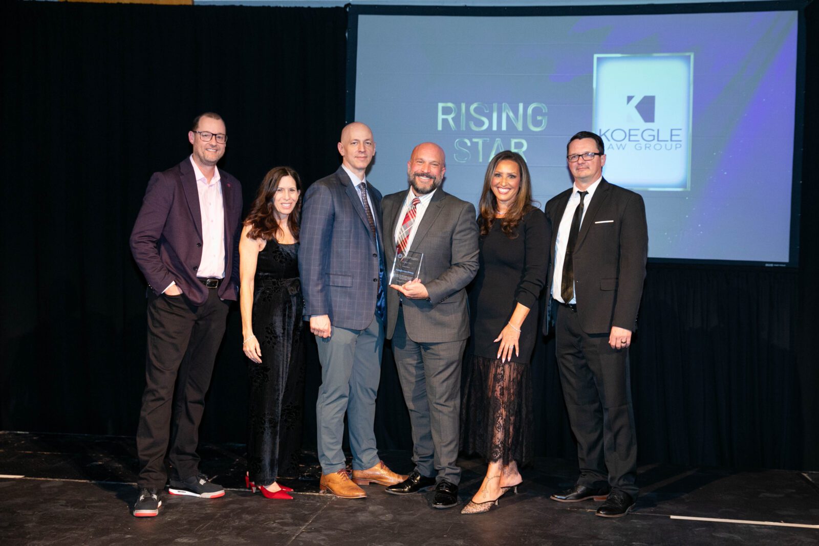 Group photo of the Koegle Law Group team on stage at an awards event with Brian Koegle holding an award in front of a screen displaying the firms logo