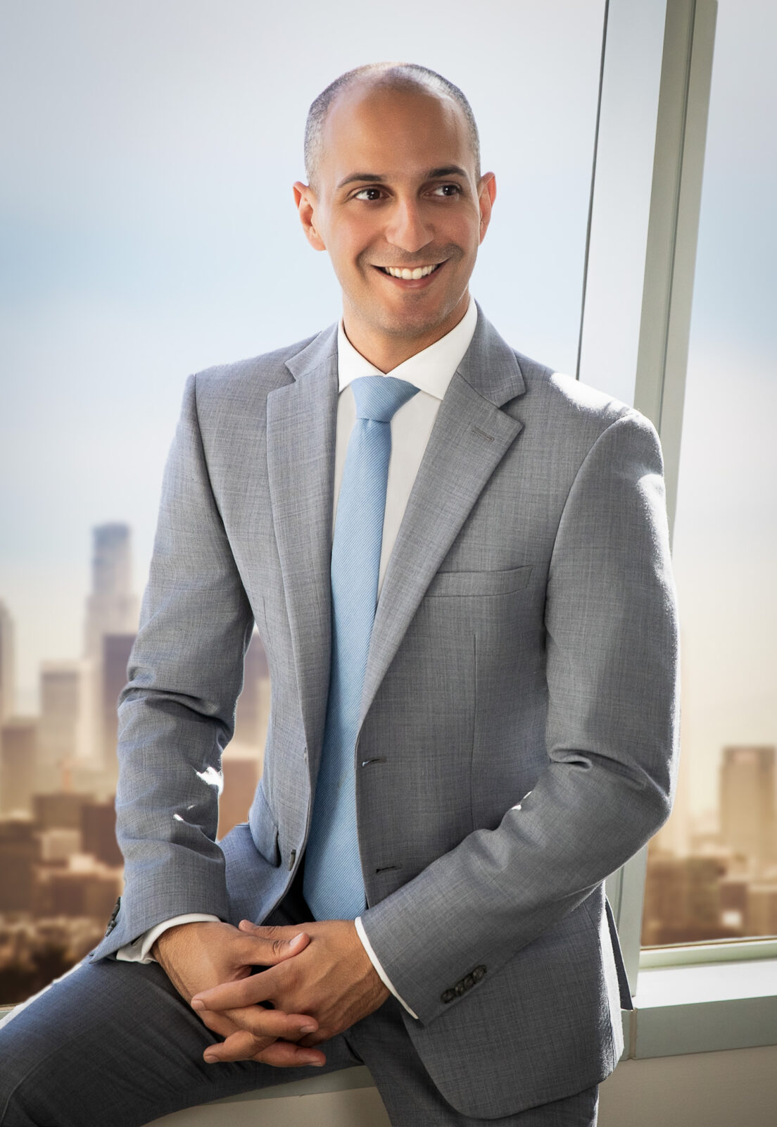 Smiling man in a gray suit and light blue tie sits by a window with a blurred city skyline in the background